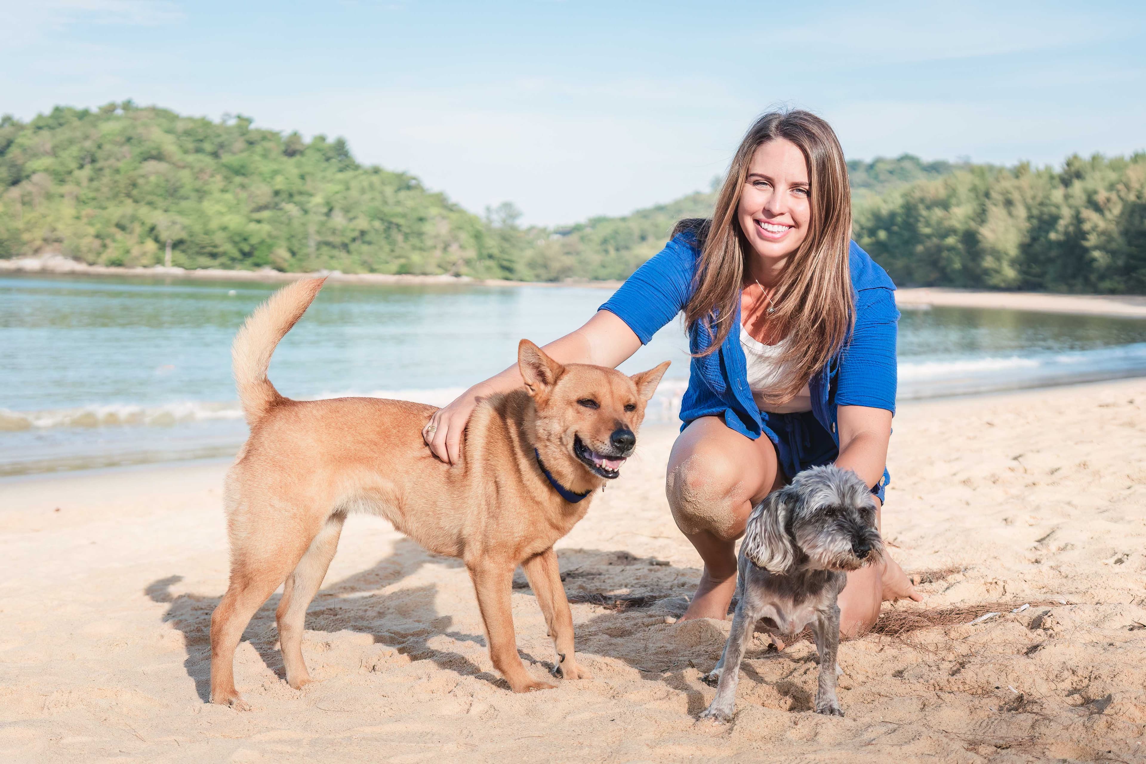 Marisa crouching on a sandy beach with her two rescue dogs, Harley and Kalinda, the sea and green hills behind them.