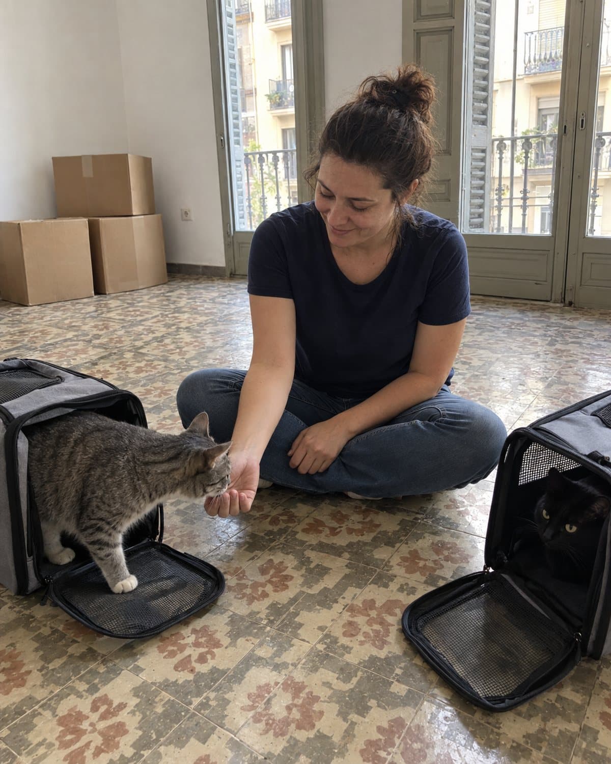 A woman sitting on the floor of an empty Barcelona apartment between two open carriers, a grey tabby stepping out to sniff her hand while a black cat watches from the other.