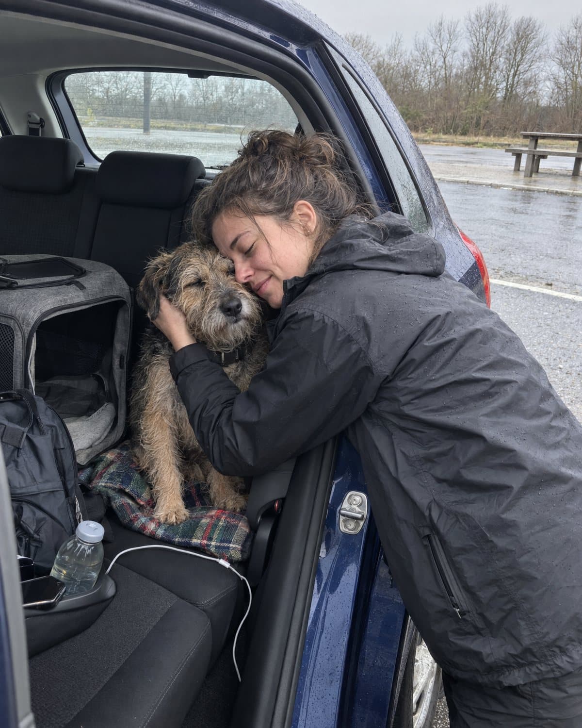 A woman leaning into the back seat of a car at a rainy French rest stop, forehead pressed to her scruffy terrier beside its open carrier.